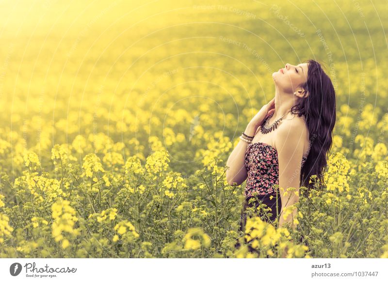 Beautiful young woman enjoying the warm sun in the nature. Attractive girl is in yellow green meadow full of flowers. With head up and eyes closed she is happy about spring or summer.
