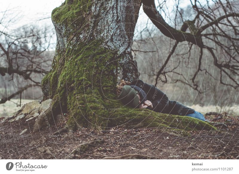 Woman lying huddled under tree with thick roots Human being Adults Life Body Hand 1 Earth Spring Tree Moss Root Tree bark Coat Scarf Cap Hair and hairstyles