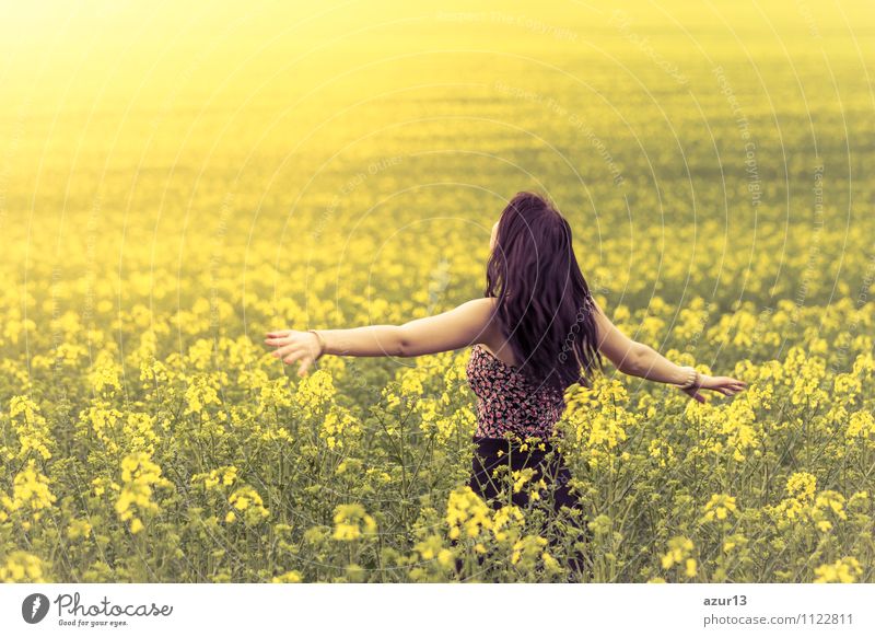 Beautiful young woman in summer on the meadow from behind with outstretched arms. Pretty girl standing in a wide field full of rape in the nature. Symbolizes happiness, health in balance and freedom full of vitality against burnout and stress.