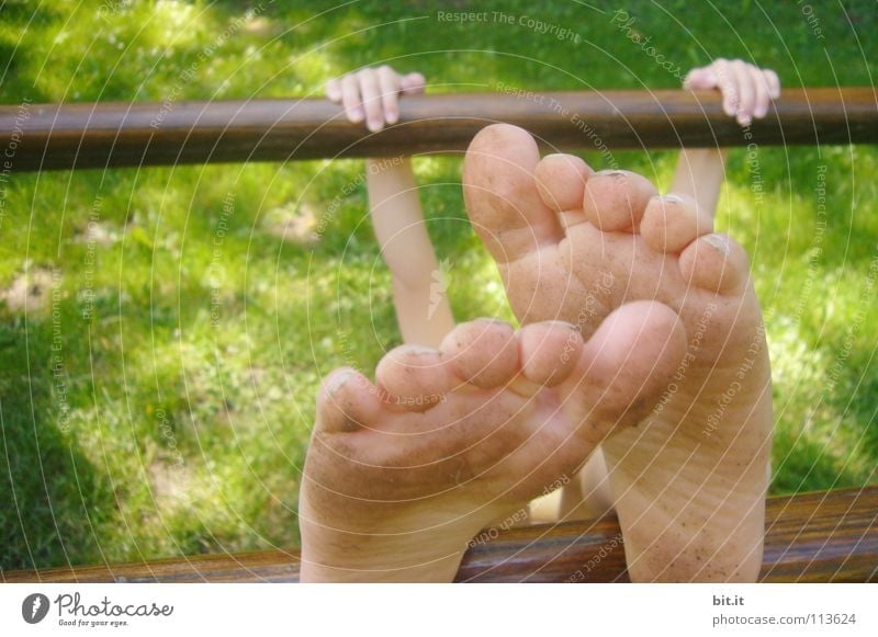 Small child hangs clamped to a wooden bar, outside in the nature. Girl is doing gymnastics on a bar, holding on with her hands and stretching her bare feet upwards. Funny feet, appear oversized and bizarre through perspective.