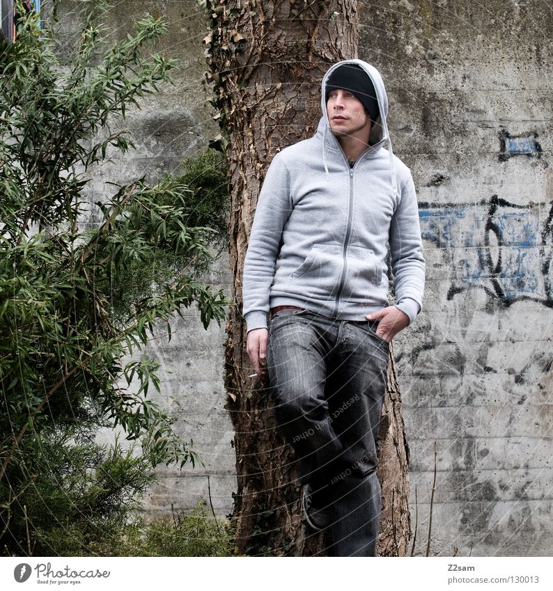 smoking break Man Youth (Young adults) Stand Smoking Cap Tree Green Bushes Style Human being Posture Hooded (clothing) tree wall Graffiti Jeans Lean Looking