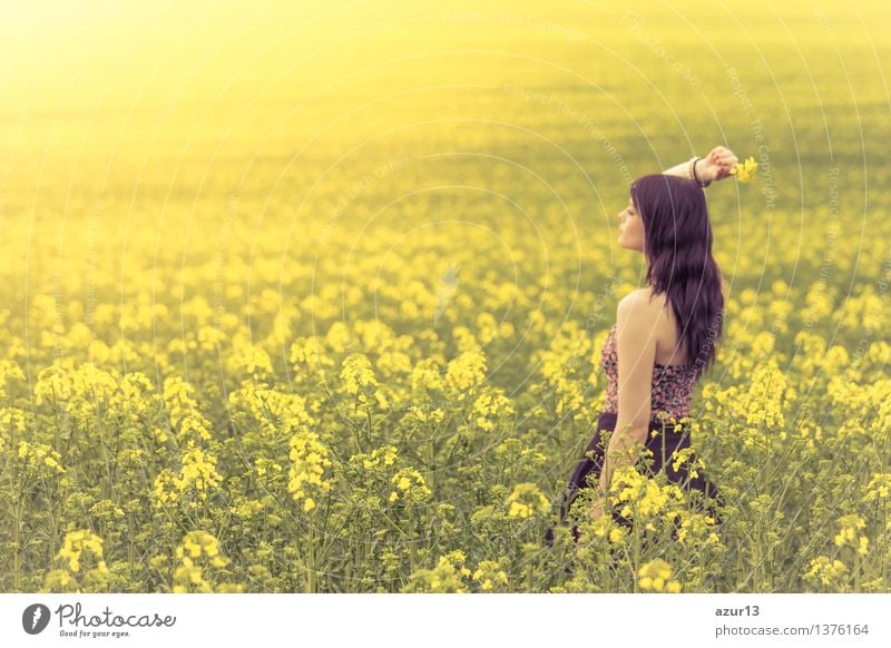 Beautiful young woman in summer on yellow meadow from rape to horizon. Pretty girl with zest for life enjoys the sunshine break and life. Rest and recharge energy from time stress in environment and nature idyll. Laterally from behind.