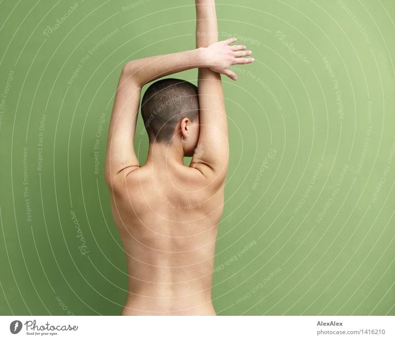young woman with very short hair stands naked with half stretched arms with her back to the camera in front of a green wall and seems to look to the right Life