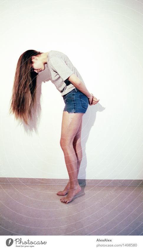 Young, athletic, slender woman stands sideways in front of a white wall with her head bent forward so that her long brunette hair hangs down in front of her face