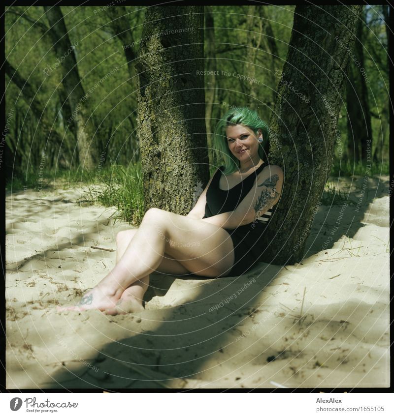 Young tattooed woman with turquoise hair sits barefoot in beach forest leaning against tree and smiles at camera Joy Relaxation Summer vacation Sunbathing