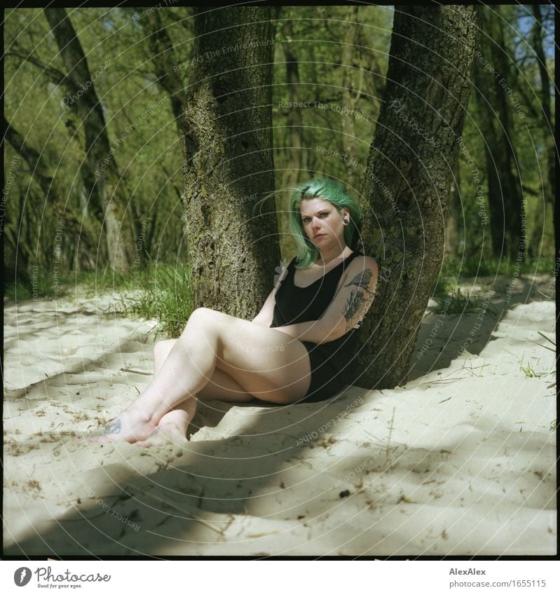 Young woman with turquoise hair sits barefoot in beach forest leaning against tree and looks skeptically at camera Calm Trip Summer vacation