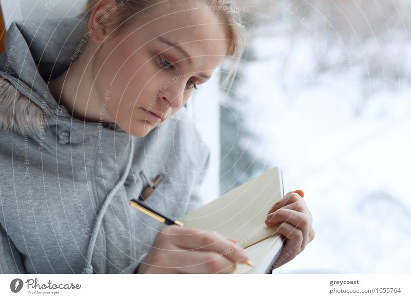 Young girl writing in her journal while sitting at a large window. Calm Reading Winter Education Student University & College student Office Girl Young woman