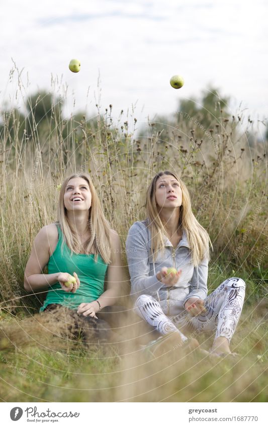 Two playful girls with long blond hair juggling outdoors as they sit side by side in long grass in the countryside Fruit Apple Happy Beautiful Wellness