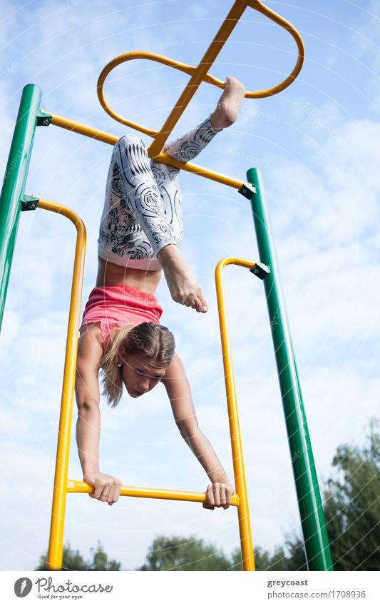 Beautiful young gymnast working out outdoors on a set of colourful metal bars hanging by her legs against a cloudy blue sky Playing Sports Child Girl Woman
