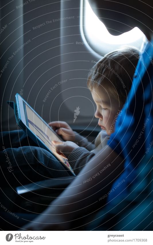 Little boy sitting in his seat during a flight playing contentedly with a tablet computer in an airplane watched by his mother Leisure and hobbies Playing