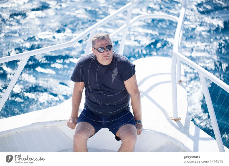 High angle view of a middle-aged man wearing sunglasses sitting on the prow of a boat on a hot summer day with the sparkling ocean below Relaxation
