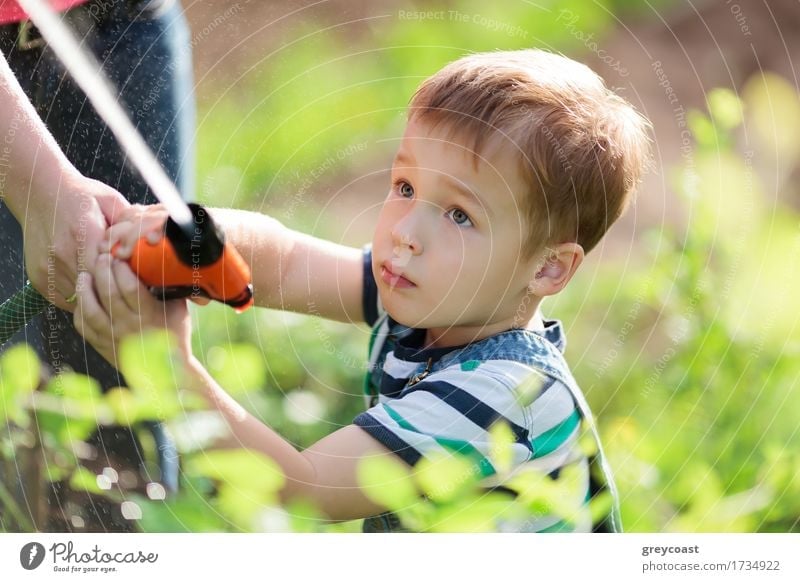 Cute serious little boy playing with a jet of water from a sprinkler being held by his mother in the garden as he stands amongst green leafy shrubs Playing