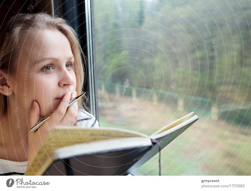 Young woman on a train writing notes in a diary or journal staring thoughtfully out of the window with her pen to her lips as she thinks of what to write.