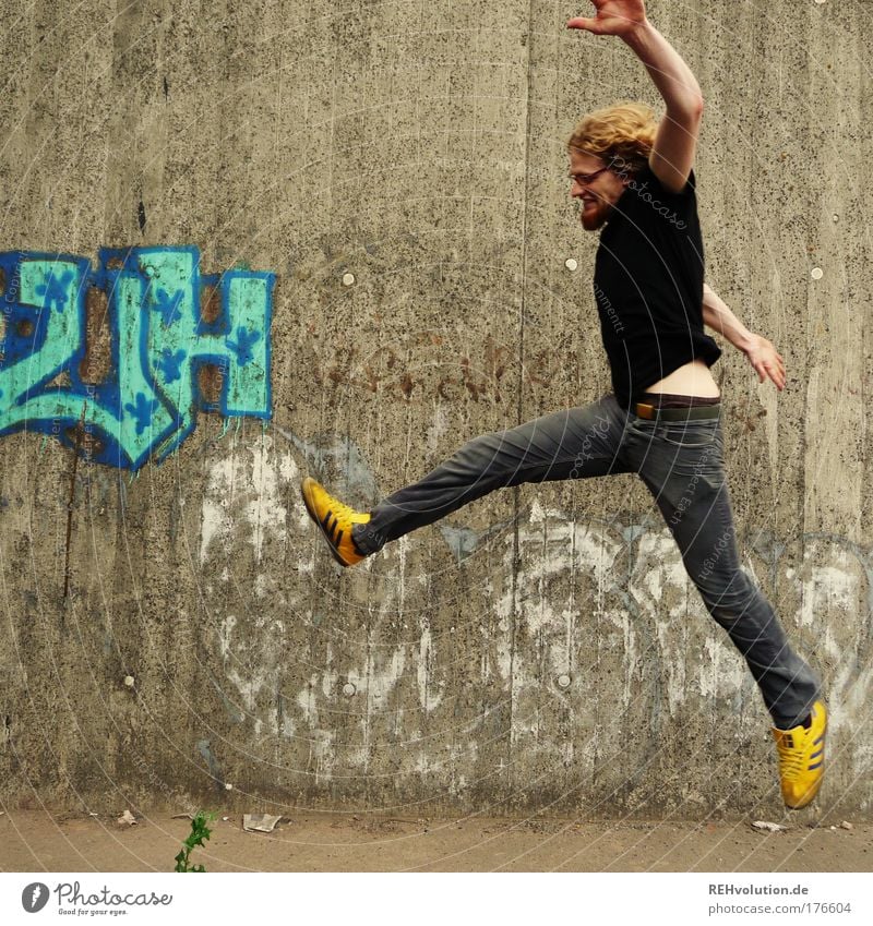 Young man jumps in front of a concrete wall Colour photo Exterior shot Copy Space left Neutral Background Day Full-length Profile Forward Style Joy Happy
