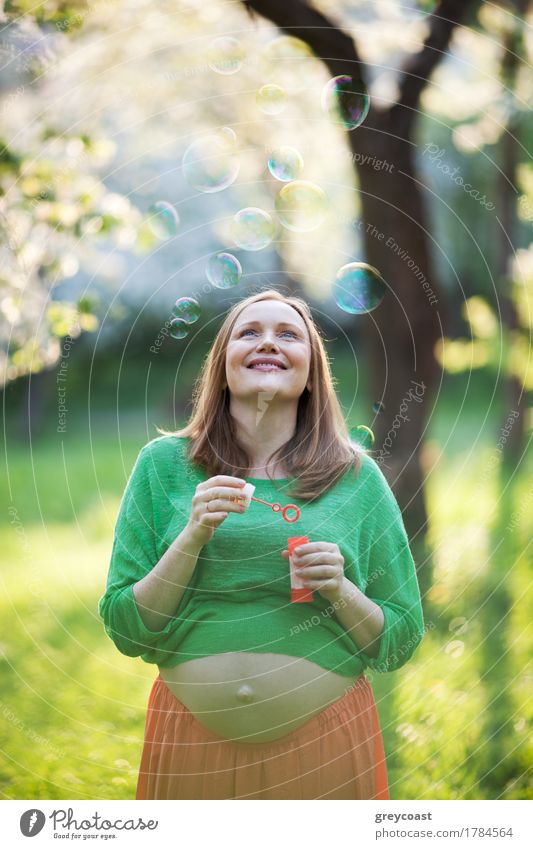 Cheerful young pregnant woman enjoying worry-free time in the park. She looking at flying bubbles on background of blurred nature scene on sunny day Joy Happy