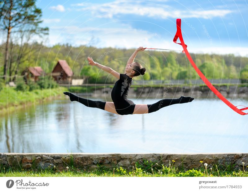 Young gymnast girl doing exercise with red ribbon outdoor. She making leg-split in a jump on background of pond and woods Happy Body Summer Sports Success Child