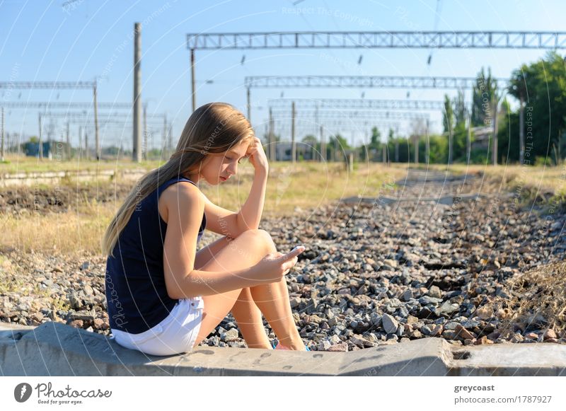 Girl teenager in top and shorts using cellphone while sitting on concrete of unfinished rail track in the countryside Summer Telephone PDA Woman Adults