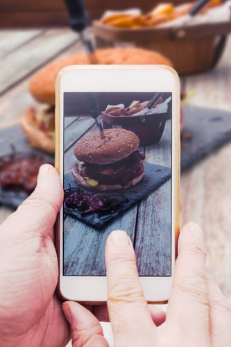 Hand with smartphone mobile phone, photo of Burger on table Food Meat Bread Roll Cheeseburger Bacon social media share Internet Table Telephone Cellphone PDA