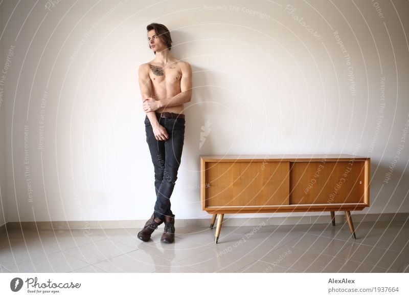 young man with bare torso and tattoo leans against the wall in a bright room with sideboard Lifestyle Style Body Contentment Furniture Room Cupboard Young man