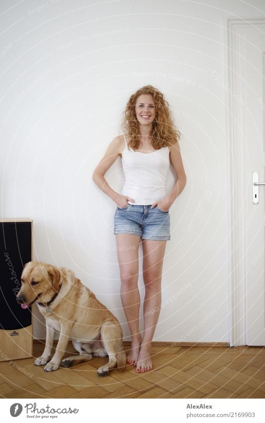 young redhead woman with curls and freckles stands barefoot in hot pants in front of white door and smiles, next to her blond labrador Lifestyle Joy pretty