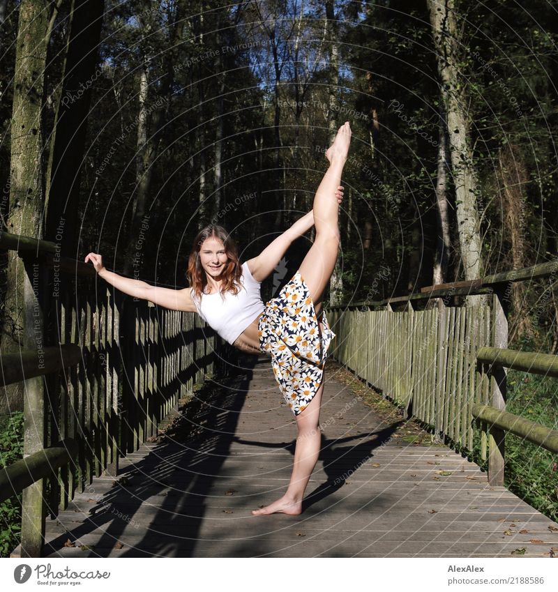Young, very athletic and muscular woman is standing on a wooden bridge in the woods doing a dance pose with one leg stretched up over her head, she is wearing a bellyless t-shirt and is barefoot