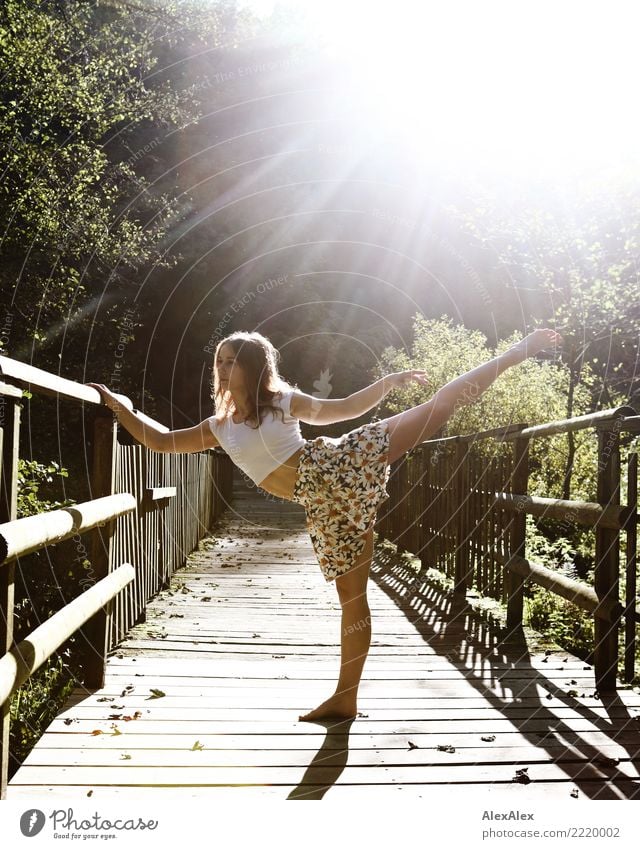 young, athletic woman stands barefoot and in the backlight on a wooden bridge in the forest and holds one leg stretched and straight up in a dancer's pose Joy