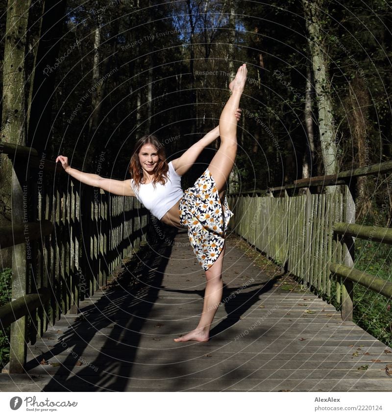Young, very athletic woman with six pack stands barefoot on a wooden bridge in the forest and holds one leg stretched over her head pretty Athletic Fitness