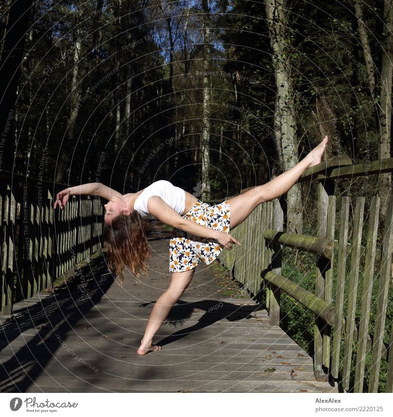 Young, very athletic and slim woman does a dance stretch on the grounds of a wooden bridge in the forest, she is barefoot and leans one leg on the railing, the head all the way back