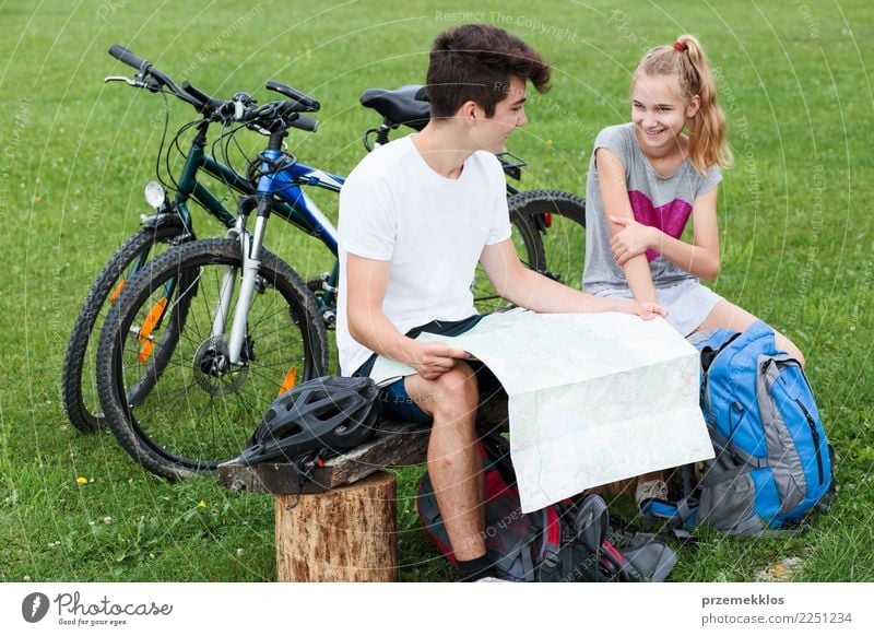 Young smiling boy and girl having a break sitting on bench during bike trip and planning a further part of trip with map equipped with helmets backpacks and bicycles