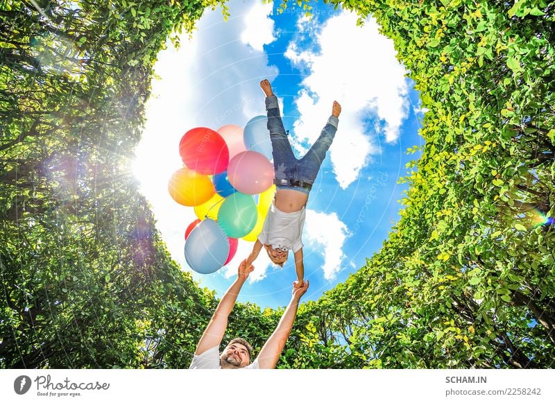 Boy flying with balloons. Dad circling his son high above his own head. All around are high green bushes. Clear blue sky in background. Bottom view. Lifestyle