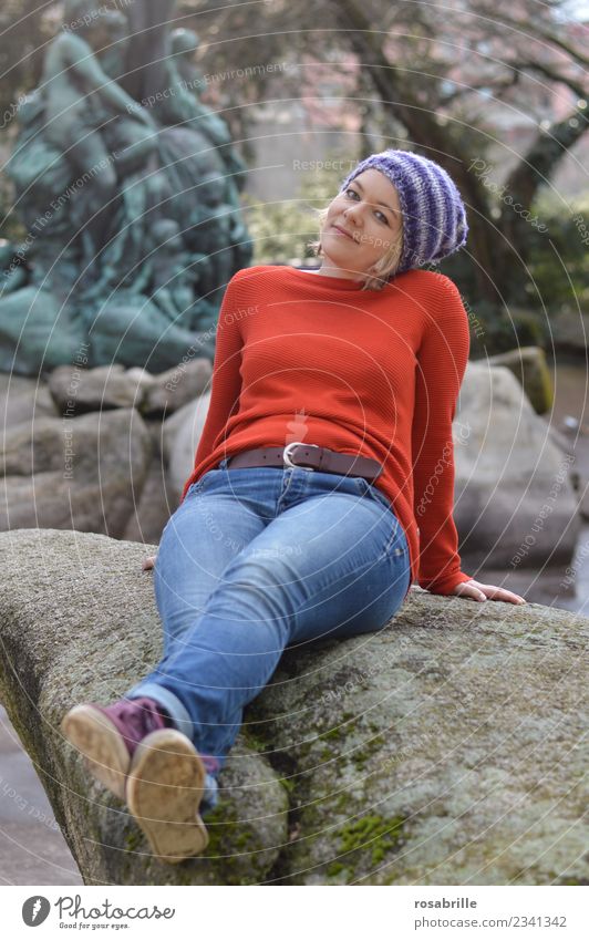 young blonde woman with purple knitted hat sits in her free time relaxed and expectant in a park on a stone and puts her head slightly tilted Joy Well-being