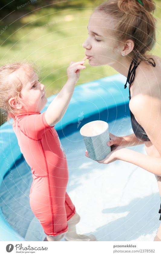 Teenage girl with her little sister spending time together in the swimming pool in a garden enjoy eating ice cream on a summer sunny day. Family quality time