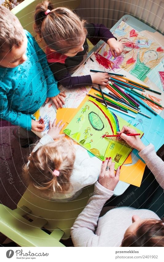 Mom with her little daughters and son together drawing a colorful picture of playing children using pencil crayons sitting at table. Photo from above Lifestyle