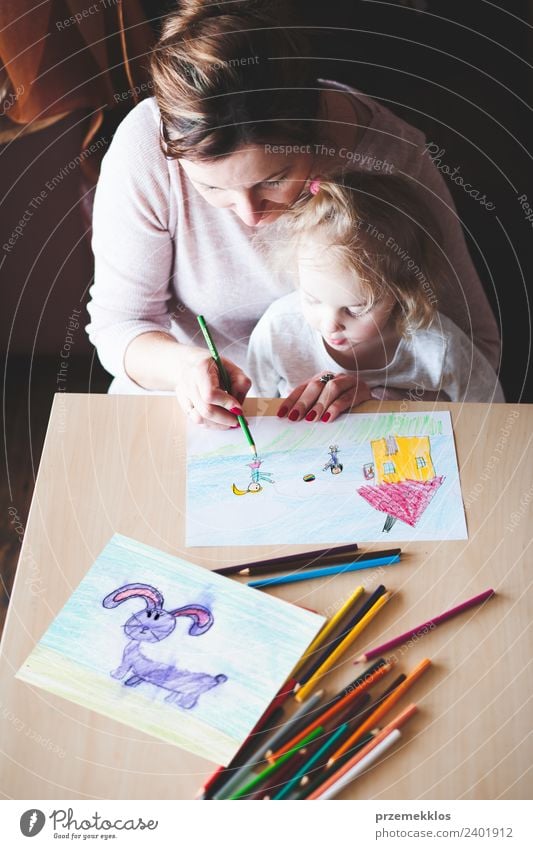 Mom with little daughter drawing a colorful pictures of house and playing children using pencil crayons sitting at table indoors. Shot from above Lifestyle Joy