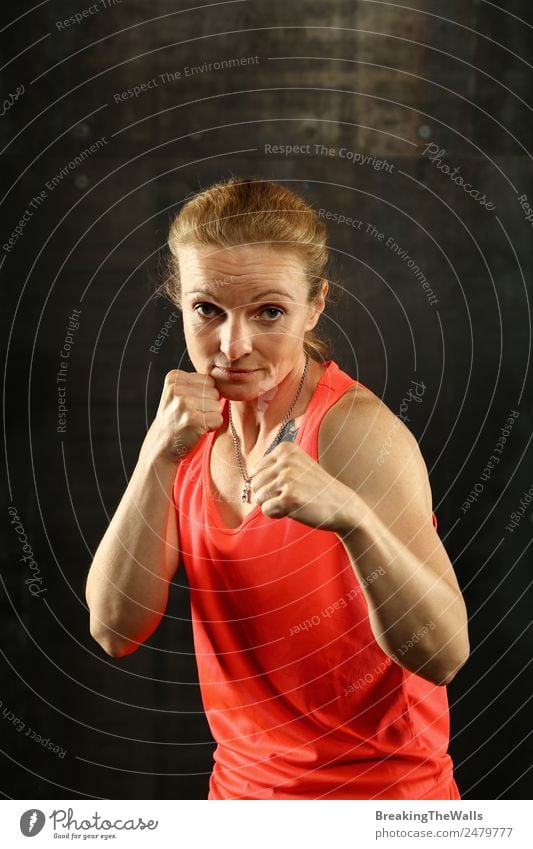 Close up front portrait of one young middle age athletic woman in sportswear in gym over dark background, standing in boxing stance with hands and fists, looking at camera