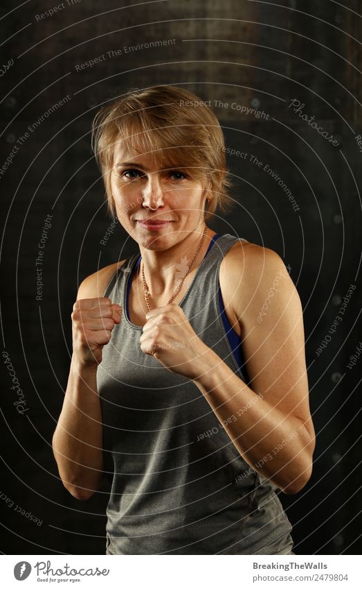 Close up front portrait of one young middle age athletic woman in sportswear in gym over dark background, standing in boxing stance with hands and fists, looking at camera