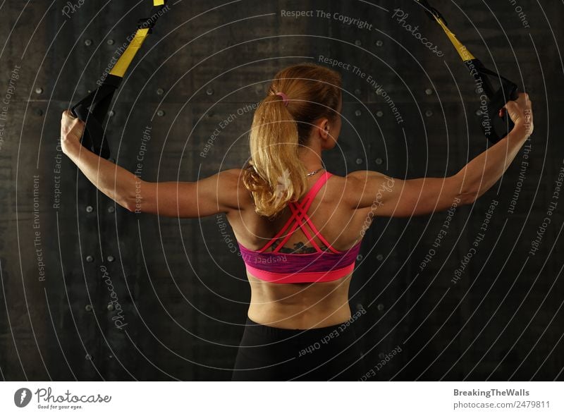 Rear view portrait of one young middle age athletic woman at crossfit training, exercising with trx suspension fitness straps over dark background Lifestyle