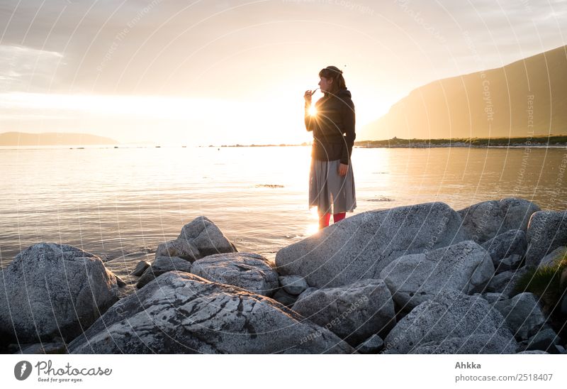 Young woman with pipe standing on stones on the shore of the North Sea in front of the midnight sun Midnight sun Back-light Sun bank Fjord Rock Ocean Light