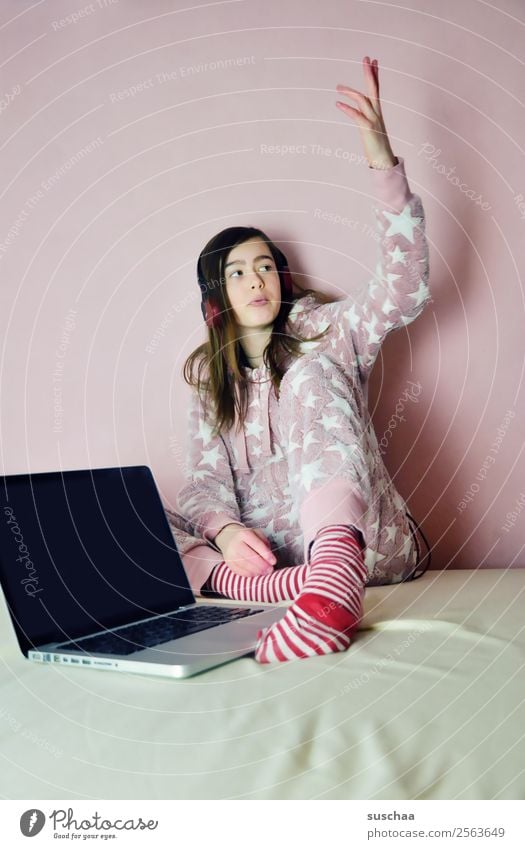 teenager with headphones sits on her bed in front of a pink wall with a laptop in front of her and lifts one arm up girl Child Young woman 13 - 18 years