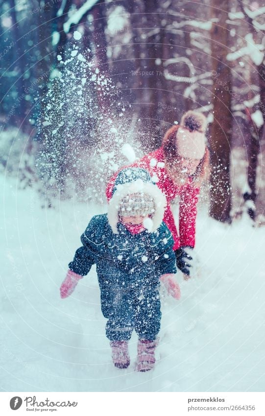 Mother is playing with her little daughter outdoors on wintery day. Woman is throwing snow on her child. Family spending time together enjoying wintertime. Woman is wearing red coat and wool cap, toddler is wearing dark blue snowsuit