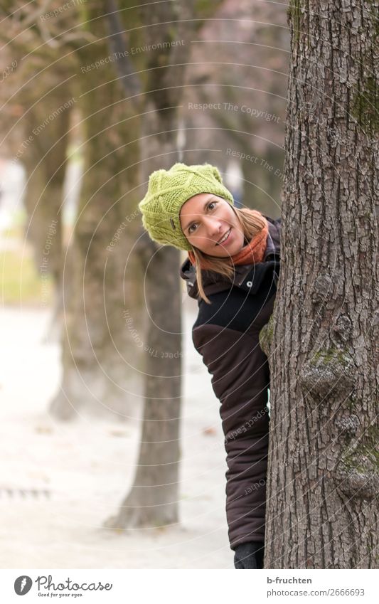 Woman looks out from behind a tree Adults Face 1 Human being Autumn Tree Park Coat Scarf Cap Brunette Observe Touch Hang Communicate Smiling Looking Playing