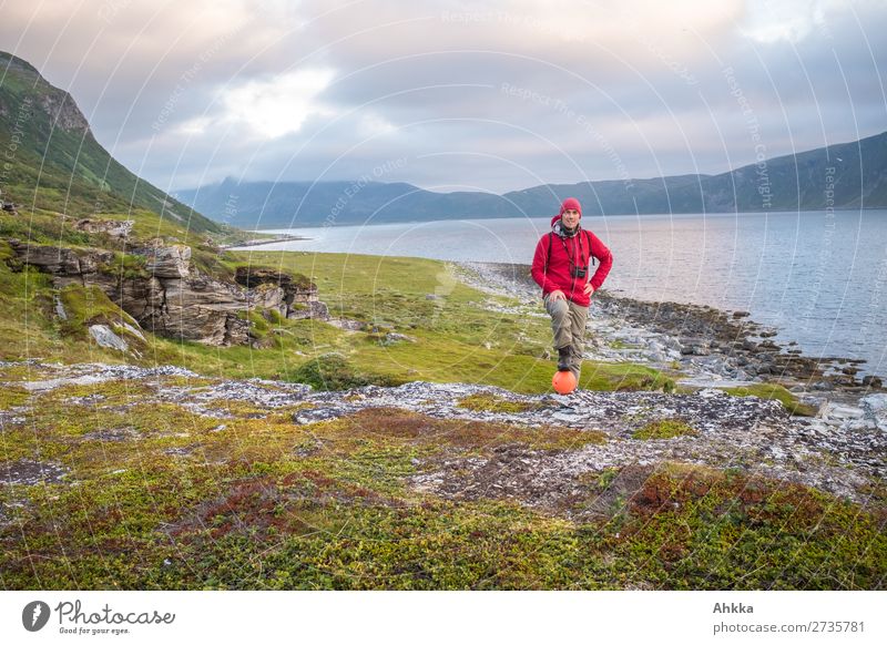 Young man with binoculars posing by fjord in evening light with view of sea and mountains Red Fjord pose Binoculars Green Pride Research Curiosity Freedom