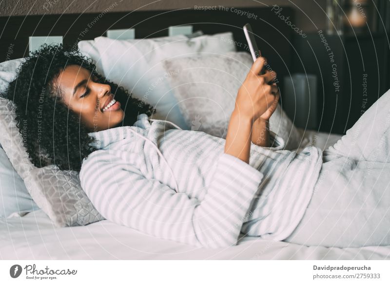 close up of a pretty black woman with curly hair smiling and using phone on bed looking away Woman Bed Portrait photograph Close-up PDA Telephone Mobile