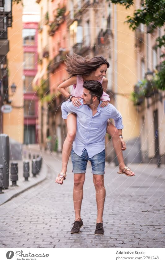 Happy girlfriend jumping on boyfriend's back in the street Couple piggy back Jump Movement Cheerful Smiling Joy Emotions Vertical Day Building Stand Beautiful