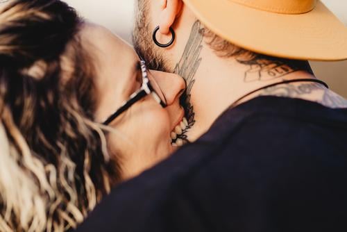 Young lady biting guy in tattoos Couple Tattoo Neck Eyeglasses Earring snapback Lady Guy Youth (Young adults) Hipster Man Woman embracing romantic Embrace Art