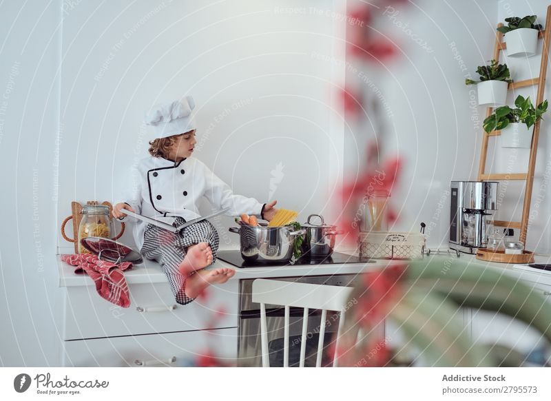 Boy in cook hat with book sitting near pots on electric fryer in kitchen Cook Boy (child) Book Pot Kitchen chef Child Vegetable Hat Volume Reading Stove & Oven