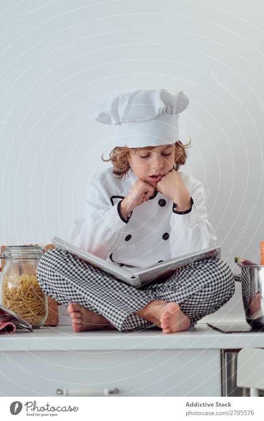 Boy in cook hat with book sitting near pots on electric fryer in kitchen Cook Boy (child) Book Pot Kitchen chef Child Vegetable Hat Volume Reading Stove & Oven
