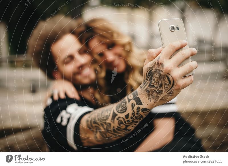 Couple making selfie via smartphone Selfie PDA using Technology focus Smiling Embrace Sit Exterior shot Arm Hand Tattoo Alternative Solar cell Mobile Day