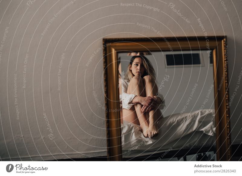 The reflection of female embracing her knees sitting on a bed. Horizontal indoors shot. woman mirror looking away morning home pensive sleepy thoughtful