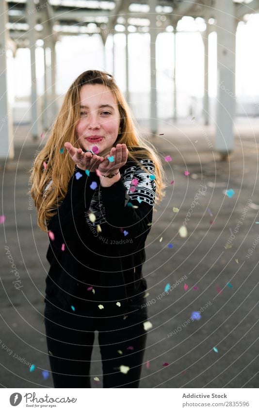 Pretty young girl blowing the confetti from her hands and looking at camera. Horizontal outdoors shot. woman fun colorful bright pretty standing flying hair